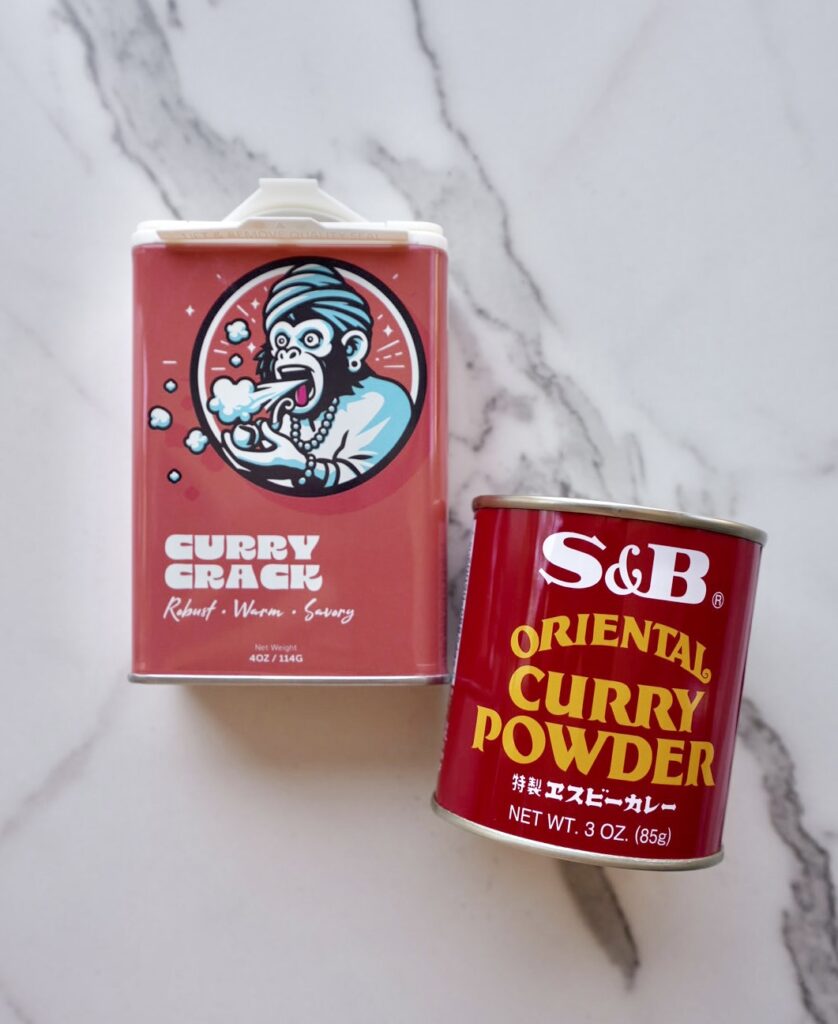 Two containers of curry powder on a marble surface—one labeled "Curry Crack" with a cartoon monkey breathing fire, and the other labeled "S&B Oriental Curry Powder" in a red tin.